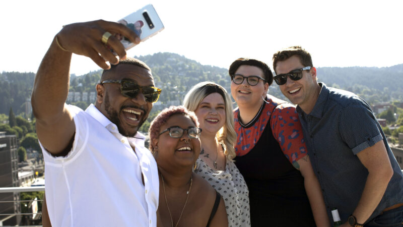 A group of friends takes a selfie on a rooftop with green hills in the background.