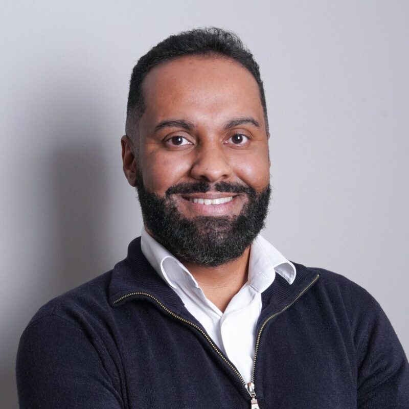 Ravi has medium skin and dark hair with a beard and bright smile. He is wearing a while button-down shirt with a navy blue quarter zip sweater.